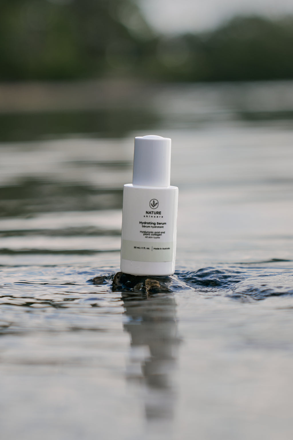 Organic Hydrating Serum bottle standing on a rock in water with blurred natural background