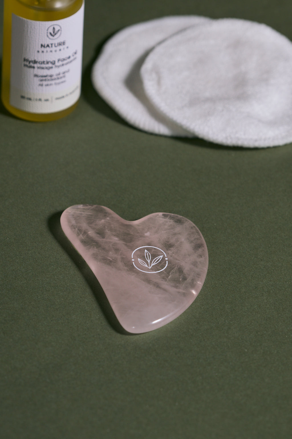 rose quartz gua sha with a bottle of skincare product on a green background